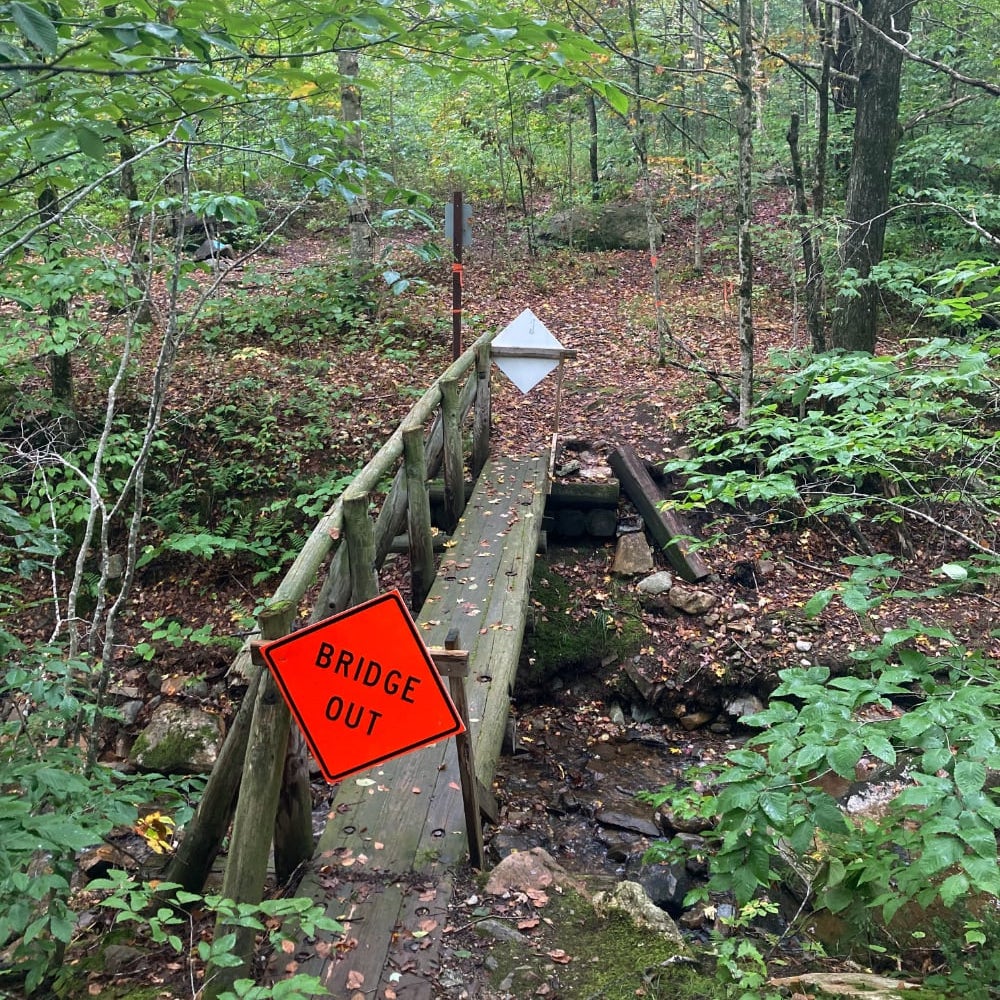 Photograph of a wooden footbridge over a stream with an orange 'bridge out' sign.